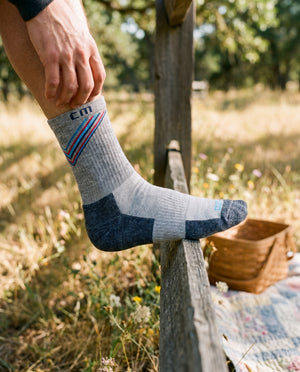 Man wearing Climatesox E-Zmate Men's Linkable Merino Wool Hiking Crew Socks with foot resting on a rustic fence rail in a sunny meadow.